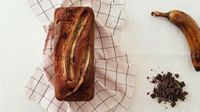 a chocolate banana bread on a kitchen towel, view from top, with a ripped banana and some dark chocolate chunks on the side
